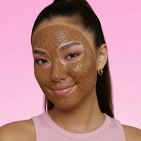 A smiling young woman wears a textured brown exfoliating facial mask against a solid pink background.